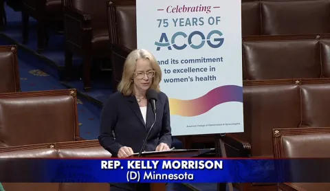 Representative Morrison stands on House floor with poster celebrating "75 years of ACOG" behind her